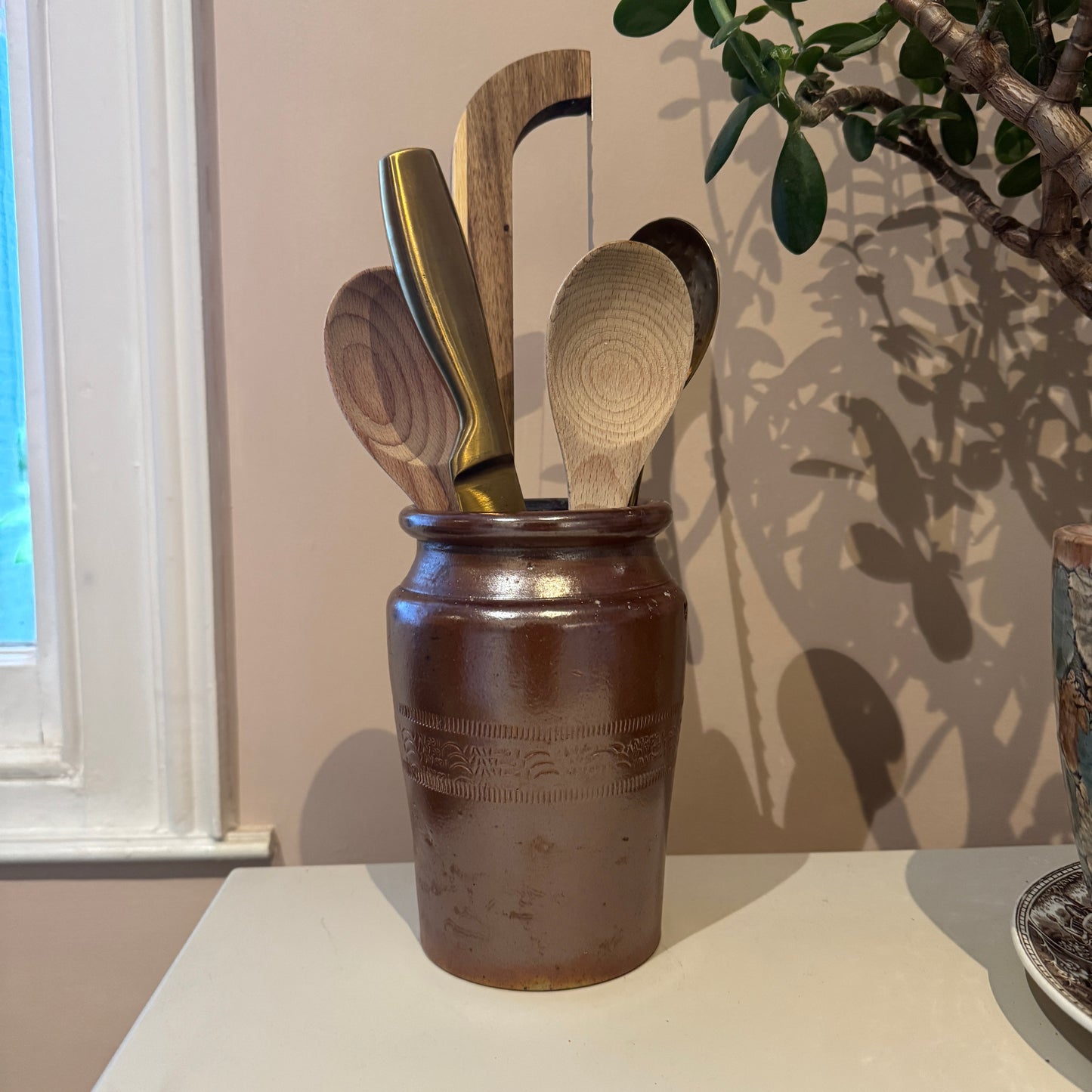 Set of wooden utensils in a brown ceramic container on a light surface with a plant in the background.